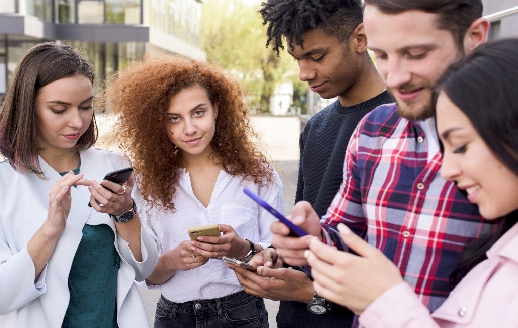 Young girl looking Camera while standing with her friends using cell phone The Negative Impacts of Social Media on Youth